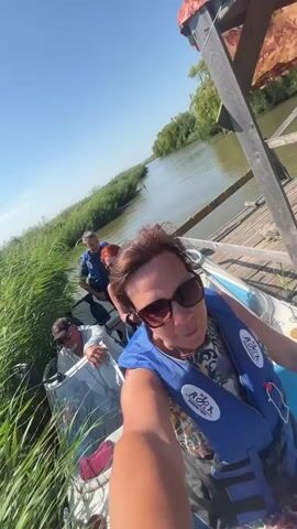 A woman in sunglasses and a blue life jacket is taking a selfie video on a boat. Three other people are visible in the boat behind her. The boat is docked next to a wooden pier. Tall green reeds line the water's edge. The sky is clear and blue.