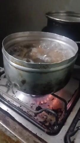 A medium-sized, well-worn silver pot is placed on a lit gas stovetop, the water inside actively boiling with visible food items. The pot is round and appears to be a standard cooking utensil, showing signs of previous use through discoloration and minor wear. The stovetop is metallic with dark, possibly burnt or stained areas around the burner. The gas flame is orange and blue, indicating combustion. In the background, out of focus, is another pot. The video's focus is on the boiling process happening inside the pot on the burner.