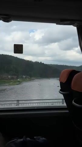 The video shows a view from a bus window of a river flowing through a forested valley. The river has several small boats or kayaks on it, and there are houses visible on the riverbank. The sky is cloudy. Two seats from the bus are visible in the foreground.