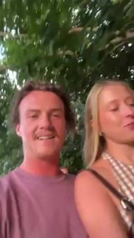 A man and a woman are standing together, smiling and laughing at the camera. The man has brown hair and is wearing a pink t-shirt. The woman has blonde hair and is wearing a striped halter top with a black purse strap over her shoulder. They are standing outside with green trees in the background. The lighting appears to be natural.