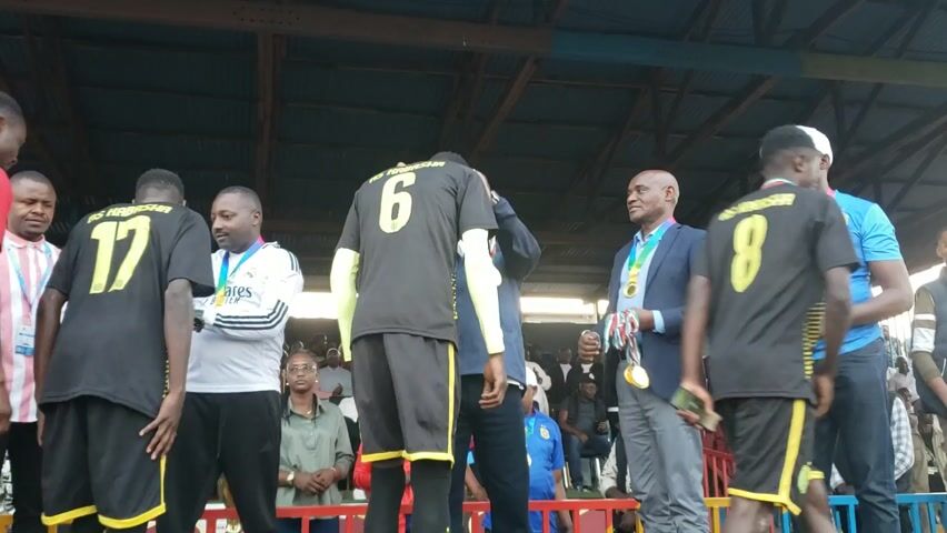 Here is a detailed description of the video:

**Scene:** The video captures an award ceremony taking place on a raised platform.

**People:** Several individuals are present, including:
*   Members of a soccer team, identified by their jerseys displaying numbers and the team name "AS Kabasha."
*   Officials or organizers, recognizable by their formal attire (suits and blazers) and the fact that they are distributing medals.
*   Spectators in the background, watching the ceremony.

**Objects:** Key objects include:

*   Medals: These are being presented to the soccer players.
*   Jerseys: The soccer players are wearing black jerseys with yellow numbers and the "AS Kabasha" team name.
*   Platform: The ceremony is taking place on a raised platform, possibly a stage or stand.

**Actions & Interactions:** The video shows:

*   Medal distribution: Officials are handing medals to the soccer players, congratulating them as they receive their awards.
*   Shaking hands: There are instances of players shaking hands with the officials as they receive their medals.
*   General interaction: People standing and observing the ceremony.

**Environment:** The scene is set outdoors, likely at a sports field. The location appears to be under some kind of shelter, possibly a simple stadium structure, as suggested by the metal roofing visible. There is a painted railing in the foreground.
