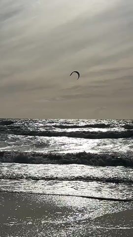 The video shows a view of a choppy ocean with waves crashing onto a sandy beach. In the sky, a kite surfer is visible. The sun is low in the sky, casting a bright reflection on the water. The overall atmosphere is serene and natural.