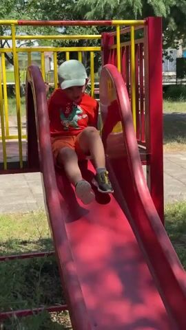 A young child wearing a light blue baseball cap, an orange t-shirt with a dinosaur graphic, and orange shorts is sliding down a red plastic slide. The child is wearing dark shoes with yellow accents. The slide is located outdoors, with grass and trees visible in the background. The child appears to be on the lower portion of the slide, with a pile of pinecones gathered at the bottom. The child's body is angled to the right of the frame, with their legs extended and their arms reaching towards the slide. The child's mouth is open, suggesting they might be vocalizing.