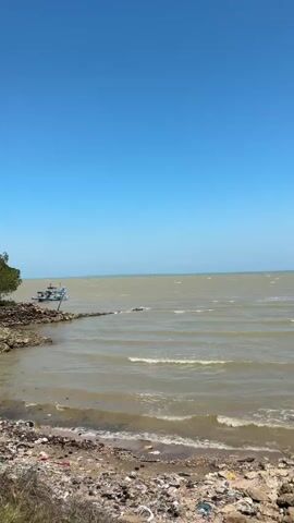 A wide shot captures a calm ocean under a clear blue sky. The water is a muted brown, with small waves lapping at a rocky, debris-strewn shore. A small blue and white boat is docked near a rocky outcrop on the left side of the frame. The shoreline is littered with various pieces of trash, including plastic bottles and wrappers. The overall impression is one of a natural environment impacted by pollution.