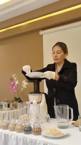 The video shows a woman using a juicer on a table covered with various ingredients. The woman is wearing white gloves and a black coat. She carefully places a plate on top of the juicer. On the table, several small jars contain different seeds, nuts, and beans. A pitcher of water is also present.  In the background, there's a wall, an orchid, and a projection screen.
