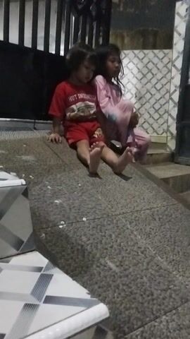 Two children are on a stone floor. One child is wearing a red shirt and shorts, and the other is wearing pink pajamas. The child in the red shirt is on their hands and knees, and the child in pink is leaning over them. The floor is wet in places. There are some white pieces of paper or debris on the floor. There is a metal railing and stairs to the left of the children. The wall behind the children has a patterned tile.