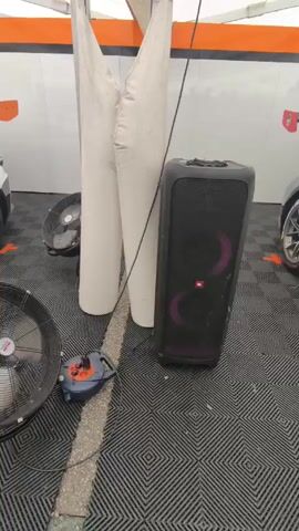 A video shows a race car team’s garage with a blue, white, and orange Porsche GT3 race car parked inside. Two men in the background are watching the team work on the car. There are two large fans and a speaker set up to keep the team cool and entertained.  A man wearing a DEKRA shirt and a camera is filming the car and the team. Other people in racing suits can be seen in the background. The garage has a black and grey checkered floor, and the walls are decorated with racing logos. There is very loud music playing.