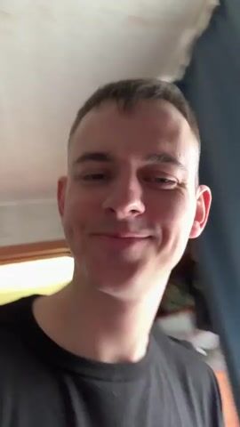 A fair-skinned man with short brown hair is holding up a book in front of his face. The book's cover features a photo of an Asian man smiling and the title "Rich Dad's Before You Quit Your Job" in English and Russian. The man in the video is wearing a black t-shirt with a white design and smiles at the camera. The background includes a light-colored wall, a partially visible curtain, and a shelf with items.