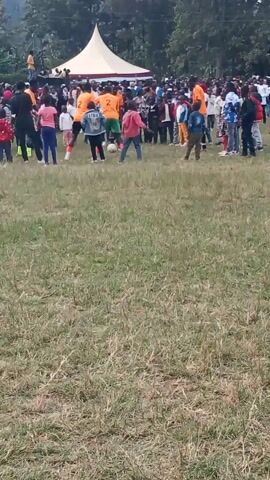 The video shows a large crowd of people gathered on a grassy field. There is a white tent or canopy in the background, and trees are visible on the far left. Some of the people are wearing brightly colored shirts, including orange. A few individuals appear to be on the field, possibly participating in an event or activity. A soccer ball is visible near the center of the frame. The overall scene suggests an outdoor gathering or event, possibly a sporting match or festival. The footage is recorded vertically, and the image is slightly blurry and shaky, indicating it was likely captured on a mobile device.