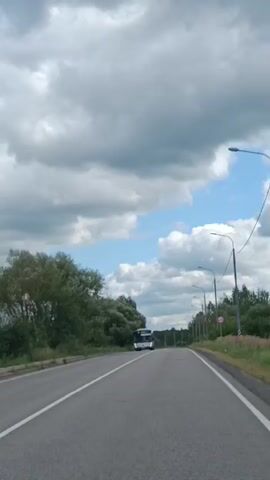 The video shows a view from a vehicle moving along a paved road. A white bus is visible in the distance traveling in the same direction. The road is bordered by green trees and vegetation on both sides. The sky is partly cloudy with patches of blue visible. The video appears to be filmed during the daytime.