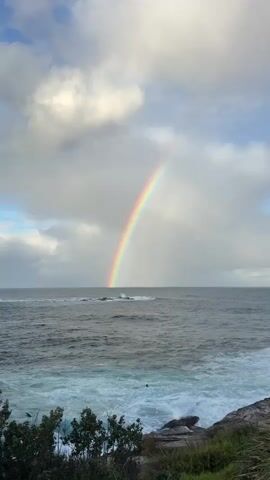 The video captures a wide shot of a turbulent ocean under a cloudy sky. A vibrant rainbow arches across the frame, its arc beginning in the distance over the water and extending upwards into the clouds. The sea is choppy, with white waves crashing against a rocky shoreline in the foreground, which is partially covered with green grass and dark shrubs.