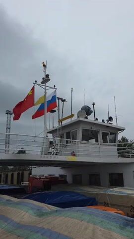 The image captures the upper section of a large white vessel, likely a ferry or cargo ship, under an overcast sky. Three national flags are prominently displayed on a mast: the flag of China (red with yellow stars) on the left, the flag of Russia (white, blue, and red horizontal stripes) on the right, and an unknown flag with vertical yellow, white, and red stripes in the center.

The vessel features a white wheelhouse or cabin on its upper deck, equipped with multiple antennas, a satellite dish, and a large loudspeaker. A metal railing surrounds this upper deck. Below the deck, tarpaulin-covered objects are visible, suggesting cargo or vehicles, including a glimpse of a red truck. In the background to the left, a tall metal tower can be seen.

The sky is cloudy and grey, indicating overcast weather conditions during daylight hours. The flags are unfurled, suggesting a light breeze.

Chinese text is visible on the side of the vessel's upper deck: "旅客定员180人", which translates to "Passenger capacity 180 people". This suggests the vessel is designed to carry passengers, possibly along with cargo. The presence of both Chinese and Russian flags strongly indicates an international border crossing or port, consistent with the location context of Nizhneleninskoe, Russia, which borders China.