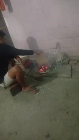 A man is seated on a wooden block with his left leg bent and his right leg extended. He is wearing a black t-shirt and white shorts. He is holding a red bucket in his left hand and is reaching towards a small fire with his right hand. The fire is in a metal grill, and there are several pieces of brick surrounding it. The ground is concrete, and there are some scattered debris. The background is a wall with pink tiles.