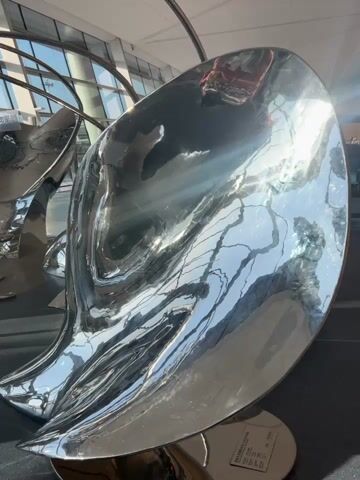 The video shows a series of highly polished, abstract, silver sculptures that appear to be made of metal. The sculptures have fluid, organic shapes, and they reflect their surroundings, including the sky, windows, and other sculptures. In the background, large windows reveal an airport terminal environment with an airplane visible outside on the tarmac. The lighting suggests it is daytime, with sunlight glinting off the sculptures and the windows. There are no people visible in the video. The sculptures are arranged in a way that suggests they might be seating or a decorative installation within the airport.