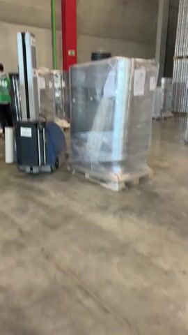 The video captures an indoor industrial warehouse in Lupanovo, Russia, during what appears to be daytime. The central focus is a robotic pallet wrapper, colored blue and silver, actively stretch-wrapping a pallet loaded with cylindrical items, likely metal cans or similar products. The items are already largely covered in clear plastic film as the machine moves around the pallet. The brand name "BREXO" is visible on the robot's casing and its upright section.

In the background, numerous other pallets with similarly wrapped goods are visible, along with tall stacks of products that resemble canned items. The warehouse features a concrete floor, high ceilings, and concrete walls or pillars. A red pillar with a red emergency sign and a green pipe are also present in the background. At the beginning of the video, a person wearing a high-visibility vest, with the number "16" partially visible, walks past in the foreground. Paper labels are affixed to the wrapped pallets, but their text is not discernible. The scene illustrates an automated packaging operation within a large storage facility.