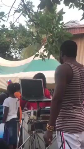 The video shows a gathering of people, possibly at a celebration or event, indicated by a tent and what appears to be a DJ setup with a laptop and speakers. The atmosphere seems lively, with music playing in the background. A man in a striped tank top and shorts is visible in the foreground. Other people, including children, are scattered around the space. The setting appears to be outdoors with foliage and trees visible in the background.