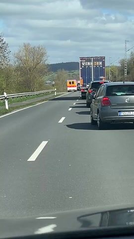 The video shows a highway with stopped traffic. In the distance, an ambulance is visible. A large truck with the name "VERDIJK EUROPE" on the side is directly in front of the camera's viewpoint. Several cars are also stopped in the same lane. The weather appears sunny with a partly cloudy sky. Trees and hills are visible in the background. There is a guardrail on the left side of the road.