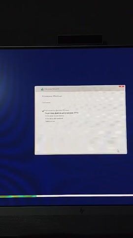 A computer monitor displays the Windows installation process. The window indicates "Installing Windows" in Russian, with the progress bar showing "Preparing files for installation (93%)". The monitor is on a desk, and the screen is dark blue behind the installation window. The computer has a camera at the top of the screen. The video appears to be taken in a domestic or office setting.