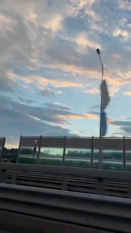 The video is taken from a moving vehicle, likely a car, traveling along a road at dusk. The sky is filled with clouds, tinged with pink and orange from the setting sun, against a backdrop of blue sky. In the mid-ground, there is a sound barrier with reflective panels that show a distorted reflection of trees and sky. A lamppost stands tall to the left of the frame. The foreground shows the road surface with white markings.