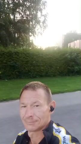 The video begins with a close-up of a man's head and shoulders as he walks. He is wearing a black jacket with yellow and white trim and earbuds. He looks to his left and then turns his head forward. The background shows a paved path, green grass, and trees. The camera then pans to the right, showing more trees and a street. The video continues to pan and move, showing a crosswalk, a lamppost, a building with a blue roof, and a tall, brick structure that appears to be a water tower. The sky is clear and a soft yellow light suggests it is either sunrise or sunset. The movement of the camera indicates the video is likely being recorded from a moving vehicle or while the person is walking.