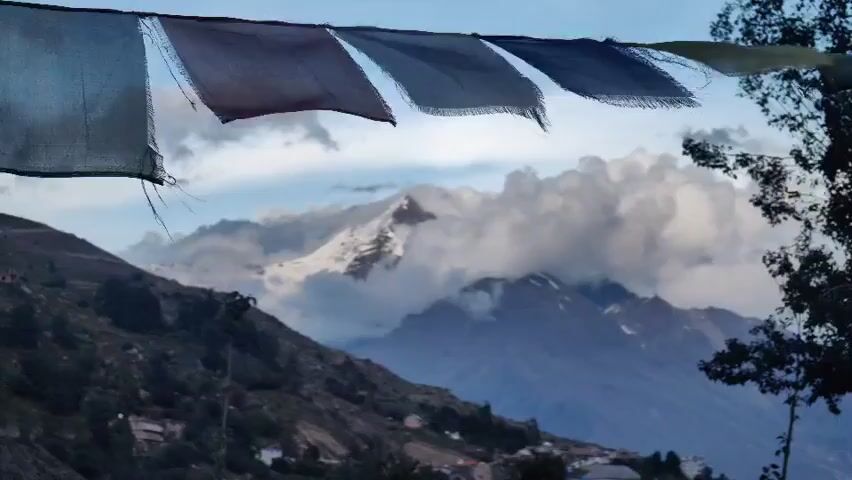 The video shows a wide shot of a mountain range with prayer flags in the foreground. The prayer flags are colorful and appear to be made of cloth, fluttering in the wind. The mountains are majestic and snow-capped, with some clouds obscuring the peaks. The sky is a clear blue with some scattered white clouds. There are some small buildings or houses visible on the slopes of the mountains, suggesting a settlement or village. The overall scene is serene and picturesque, evoking a sense of peace and spirituality.