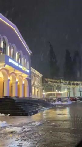 The video shows a building at night with snow falling. The building is lit up with yellow lights and has a sign that reads "ORTAZU GAME 2026 HOTEL". There are stairs leading up to the building and a wet sidewalk in front of it. There are also some trees in the background with string lights hanging between them. There are cars parked on the street. The sky is dark and the snow is falling heavily.