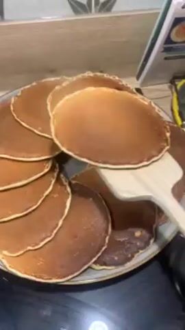 A plate piled high with golden brown pancakes sits on a reflective black surface, possibly a stovetop. The pancakes are arranged in a circular pattern, slightly overlapping one another. A wooden spatula rests on top of the pancakes, partially obscuring some of them. In the background, a wall with a decorative design and a partially visible instructional guide, possibly a recipe book, are present. The lighting is indoor and appears to be fairly bright, highlighting the texture of the pancakes.