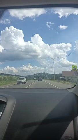 A dashboard view from inside a vehicle traveling on a road. The road has two lanes in the direction of travel. Several cars are visible on the road ahead. On the right side of the road, there are streetlights and buildings. In the distance, there is a hill with structures on top. The sky is blue with large, white clouds. The timestamp indicates the video begins at 00:00 and ends at 00:04.