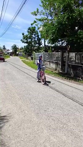 A young woman wearing a blue hijab and a light blue and white outfit rides a pink bicycle down a paved road lined with fences and trees under a bright blue sky. The bicycle is a folding bike. She is pedaling and looking ahead. In one shot, she turns and waves her hand towards the camera. Another shot shows her looking to her left and adjusting her hijab. The road appears to be in a rural or suburban area.
