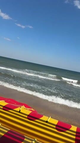 The video shows a beach scene on a sunny day. The camera is angled downwards towards the water, with the edge of a bright yellow and red inflatable object in the foreground. The ocean is calm with gentle waves breaking on the sandy shore. In the distance, two people are visible in the water, appearing to be swimming. The sky is a clear blue with a few scattered white clouds.