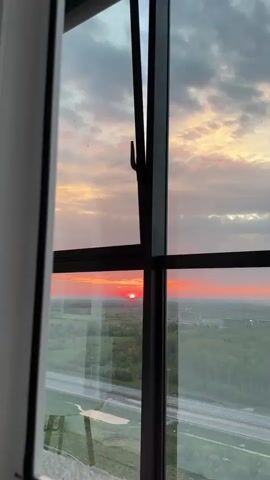The view from a high-rise window reveals a vivid sunset with a striking red sun on the horizon. A highway is visible below with sparse traffic. A green area with trees is positioned between the highway and the horizon. The sky is filled with a mix of dark gray clouds, as well as clouds colored pink and yellow by the sunset. The window has a black frame and is slightly ajar, with the handle visible. The interior of the building is visible on the left side of the frame.