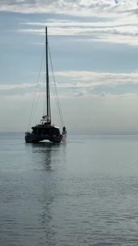 A large catamaran sailboat is captured moving away from the camera on calm, shimmering water under a partly cloudy sky. The boat features a tall mast and rigging, with a small flag visible near the top of the mast. While rigged for sailing, no sails are deployed, suggesting it might be motoring. Two individuals are discernible on the stern of the vessel. The lighting is soft and diffused, hinting at either a tranquil morning or late afternoon, creating a serene and peaceful ambiance. The boat leaves a gentle wake as it glides across the water. This scene is set in Divnomorskoe, Russia. No visible text is present in the frame.