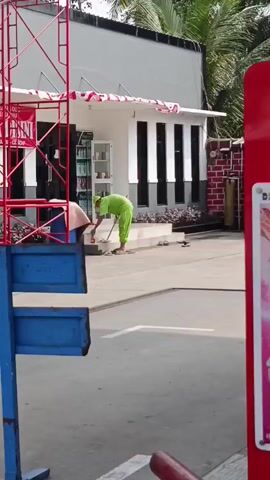 A person in a green uniform is sweeping the ground in front of a small white building with black-trimmed windows. The building has a red and white striped awning. There is scaffolding to the left of the building, and a red structure on the right. Trees are visible in the background.