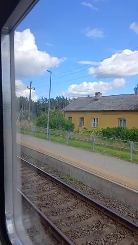 The video begins with a view from inside a moving train, looking out a window at a train station. The train tracks are visible in the foreground, with a beige platform alongside them. Behind the platform, a yellow building with a gray roof and a chimney stands under a bright blue sky dotted with fluffy white clouds. Trees and vegetation surround the building and line the horizon. A gray metal fence runs along the edge of the platform, separating it from the area behind. A lamppost is also visible on the platform. The train is in motion, indicated by the slight blur of the scenery passing by. The video is short and does not show any people or interactions.