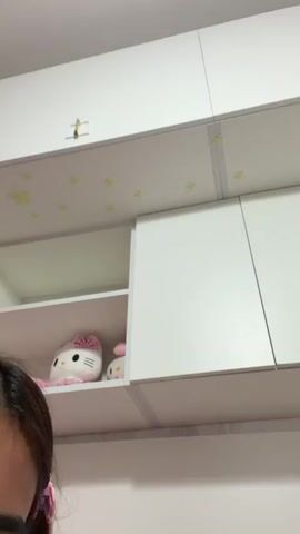 A close-up video shows a person's face and hands in the foreground, with shelves and cabinets in the background. The person has brown hair with pink clips and is looking down. Their right hand is raised, showing painted nails. The person makes a peace sign with their left hand, then moves their right hand to point towards the camera. On the shelf in the background, there are two Hello Kitty plush toys. The shelves and cabinets are white, and there are yellow star stickers on the wall above the shelves.