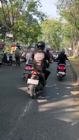 The video shows a street scene with several people riding motorcycles. On the left side of the street, there are stalls selling various items. The stalls are set up along the side of the road, and several motorcycles are parked nearby. In the center of the road, a group of people on motorcycles are riding away from the camera. The motorcycles are of various colors and models, and the riders are wearing helmets. The environment appears to be a public area with trees and buildings in the background. The lighting suggests it is daytime.