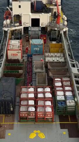 A high-angle aerial shot shows the deck of a ship loaded with cargo. The ship is at sea, with dark water visible around it. The deck is filled with various containers and equipment, including shipping containers, open-top bins, and what appear to be crates of supplies. Two individuals in orange safety suits are visible on the deck, one near the center and another further back. They are likely workers engaged in cargo operations. The overall impression is one of a working vessel at sea, prepared for or in the process of transporting goods.