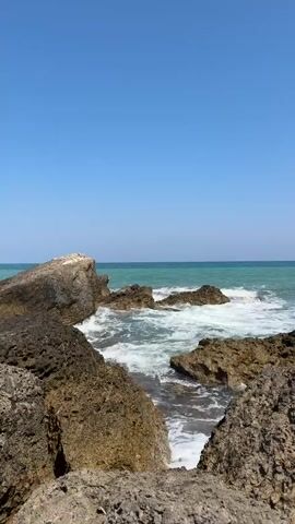 The video shows a rocky shoreline with the ocean and a clear blue sky. The waves are crashing against the rocks, creating white foam. The rocks are dark brown and grey. The water is a turquoise color. The sky is a bright, cloudless blue. The video is shot from a low angle, looking out towards the sea. The camera is stationary.