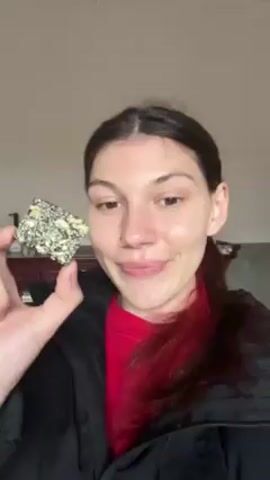 The video shows a fair-skinned woman with brown hair, partially dyed red, tied back in a ponytail, wearing a red shirt and a black jacket. She is holding a small square snack with green and white specks. She puts the snack in her mouth, pauses, and then looks directly at the camera. The background is a light-colored wall and a blurry image of furniture. The woman announces, "Let's try Chinese snack."