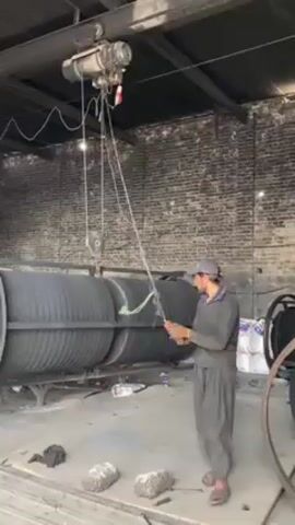 A man operates an electric hoist in a warehouse. The hoist is suspended from a beam, and its cable extends down to lift a large, cylindrical object. The man is wearing a gray outfit and a cap. The warehouse has brick walls and a metal roof structure. Other objects, including paint cans and wheel parts, are visible in the background.