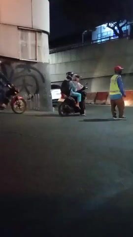 In the video, at night, several people are on motorcycles on a paved road. A person wearing a yellow vest and a red hat walks across the road. In the background, there is a concrete structure and a light pole.