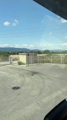 A wide shot captured from inside a vehicle shows a grey asphalt area with white parking lines. A metal gate is visible on the left, with a red object on a post in front of it. In the center, a tan colored building stands behind a fence. A red stop sign is attached to a pole in front of this building. The area is surrounded by a metal fence, and in the background, there are fields, trees, and mountains under a blue sky with scattered clouds. Electrical wires are strung across the sky. The video appears to be a short clip of the vehicle moving through the area.