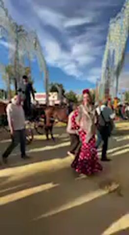 The video captures a vibrant daytime scene at what appears to be a traditional Spanish festival or feria in Jerez de la Frontera, Spain. The weather is bright and sunny, with a clear blue sky adorned with white clouds.

In the foreground, a woman dressed in a stunning, traditional red and white polka-dot flamenco dress, complete with ruffled sleeves and a long, flowing skirt, walks gracefully. She also wears a light-colored shawl and a red floral accessory in her hair, appearing to pose or interact with the camera with a joyful expression.

Around her, numerous people are visible, many dressed in formal or traditional Spanish attire. Several men are seen in suits or riding gear, some walking alongside the woman. In the background, multiple horse-drawn carriages, each with drivers and passengers, move slowly, contributing to the festive parade atmosphere. The ground is a light, sandy color, typical of fairgrounds. Large, ornate white metal archways, characteristic of Spanish ferias, frame the scene against the bright sky, adding to the grandeur of the event. No visible text is present in the video.