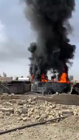 Here's a description of the video:

The video shows a scene of devastation after an apparent explosion and fire. A large plume of thick black smoke rises from a burning tank or container, with visible flames. The surrounding area is littered with debris, including broken concrete and twisted metal. Three people are visible in the video, all appearing to be men dressed in traditional Afghan clothing. They are standing amidst the rubble, observing the scene. The environment seems arid and exposed, with some distant structures visible in the background. The overall impression is one of destruction and aftermath of a violent event.