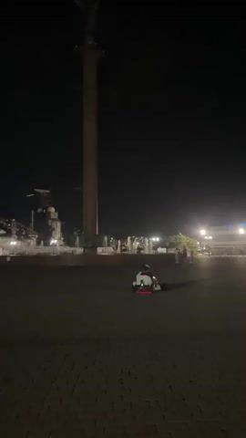 The video captures a nighttime scene in a public square. A tall monument with a statue at its apex stands prominently on the left side of the frame. In the mid-ground, a person is operating a small, white electric go-kart, which has red and white accents. The go-kart is moving across the paved square. Further in the background, there are lights from a building, and what appear to be fountains. Several other people are visible in the distance, and streetlights illuminate parts of the area. The overall atmosphere is dark, with artificial lighting creating illuminated zones.
