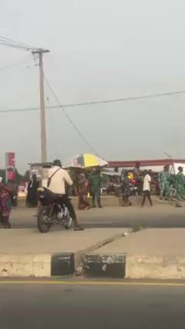 The video shows a street scene in what appears to be an urban or semi-urban environment. A man on a motorcycle is stopped on the side of the road, near a curb. Several people are standing or walking around in the background. Some are wearing traditional clothing. There is a yellow bus driving past, as well as other parked motorcycles. A sign and a fuel station are visible in the distance. A utility pole with wires stretches across the top of the frame. The overall impression is of a busy roadside area with people engaged in various activities. The sky is overcast.