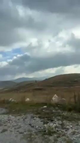 The video shows a landscape shot from a moving vehicle. The foreground is a paved road with white lines.  Beyond the road is a vast, open field of dry, brown vegetation. Low, rolling hills are visible in the distance. The sky is predominantly covered in thick, gray clouds, with some patches of blue peeking through. A roadside barrier is also seen in the view.