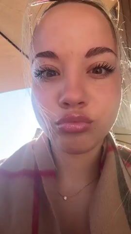 The video shows a woman with sunglasses on her head, wearing a plaid scarf and a necklace. She is at an outdoor cafe, drinking an iced coffee and enjoying a croissant. She is under an umbrella. A man adds more milk or cream to the woman's iced coffee. The sky is blue and the cafe is surrounded by trees and mountains.