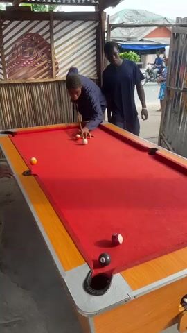 Two young Black males are engaged in a game of pool on a bright, sunny day in Badagry, Nigeria. The pool table has a vibrant red felt and a light wood-colored frame. Several billiard balls are visible on the table, including a solid orange ball, a striped purple ball with the number 9, a solid white cue ball, and a solid white ball. One of the males, wearing a dark blue outfit, is leaning over the table, holding a pool cue and appearing to be focused on his shot. The other male, also in dark blue, stands behind him, observing the game.

The setting appears to be an outdoor area, possibly a bar or recreational spot, with a thatched roof structure providing some shade. In the background, there are glimpses of buildings and people, suggesting a bustling community environment. The overall mood seems relaxed and engaged, typical of a friendly game. The lighting suggests it is daytime, with the sun casting shadows. The phrase "PREMIER" is visible on the pool table pocket.