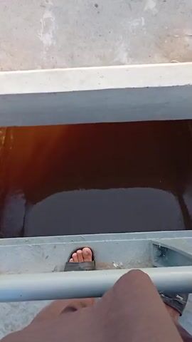 The video shows two bodies of water. First, a dark, narrow channel of water with a man standing beside it is shown from above. The man has brown skin, is wearing shorts, and is wearing a sandal on his right foot. Then, the scene cuts to a wide canal-like waterway. The water is murky and turbulent. One bank of the waterway is lined with a low, concrete barrier, followed by a concrete path, and then a grassy area, and some trees. Some debris is on the path. The water flows rapidly. The end of the path is marked by a white, low fence. The person filming is standing very close to the edge of the canal.